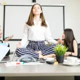 A woman meditates in lotus pose at a conference table while colleagues argue heatedly around her, creating a stark contrast between chaos and calm conversation.