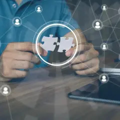Hands connect puzzle pieces, symbolizing collaboration and leading a remote team through a network of connected people icons.