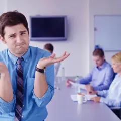 Confused man shrugs, wondering how to run meetings effectively while colleagues discuss in the background.
