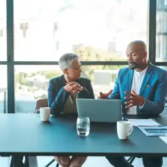 Two professionals discuss ideas for an internal communications podcast, leaning over a laptop at a conference table.