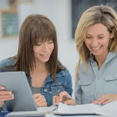 Two women smile while studying together, possibly taking general education courses online. One holds a tablet, the other a pen.