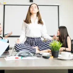 A woman meditates in lotus pose at a conference table while colleagues argue heatedly around her, creating a stark contrast between chaos and calm conversation.
