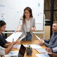 A woman leads a delegation training session, handing out papers to her team during a meeting.