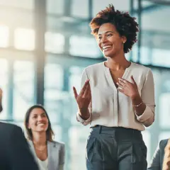 A woman leads a manager training session, engaging her audience with expressive gestures and a bright smile.