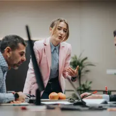 A woman in a pink blazer leads a discussion, demonstrating effective leadership and mentoring to her colleagues at a conference table.