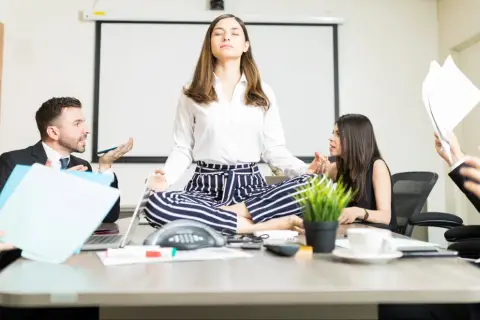 A woman meditates in lotus pose at a conference table while colleagues argue heatedly around her, creating a stark contrast between chaos and calm conversation.