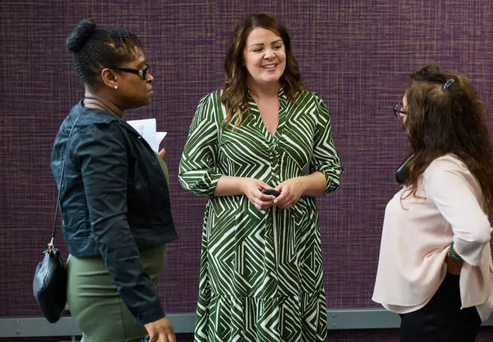Three women in conversation, one in a green patterned dress, discussing strategies for driving performance.