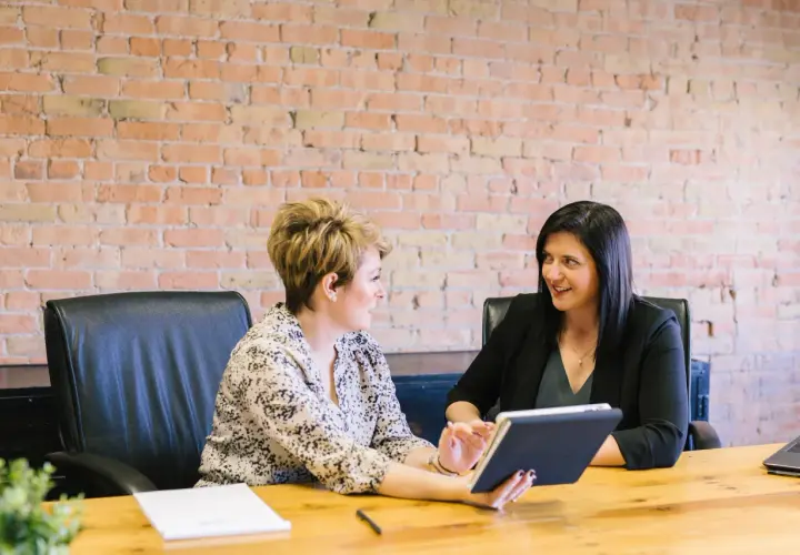 Two women in a meeting, one holding a tablet, with a brick wall background.