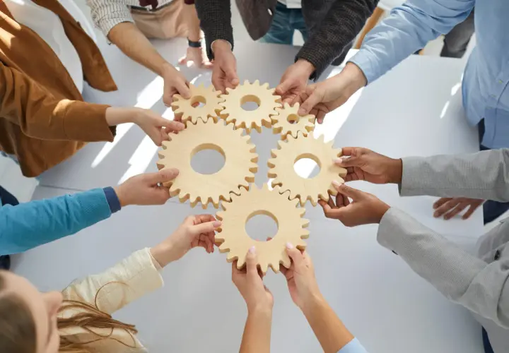 Hands connect wooden gears, symbolizing teamwork and collaboration training. Each piece fits perfectly, showing how individuals work together.