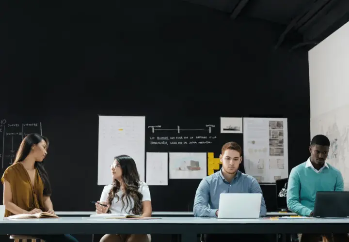 Four diverse individuals engage in a front line manager training session, collaborating around a table with laptops and notebooks.