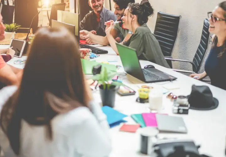 Millennials in the workplace collaborate around a table, sharing ideas and laughter. Laptops, tablets, and colorful notebooks suggest a dynamic, modern office environment.