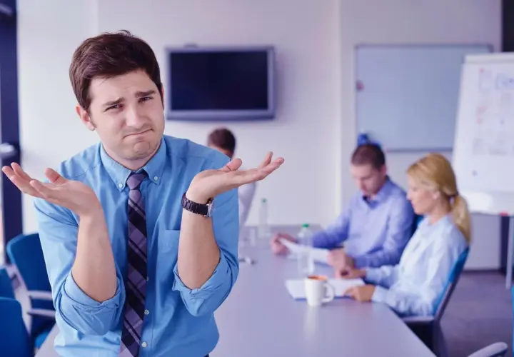 Confused man shrugs, wondering how to run meetings effectively while colleagues discuss in the background.