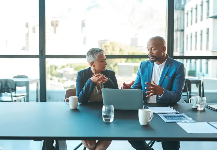 Two professionals discuss ideas for an internal communications podcast, leaning over a laptop at a conference table.