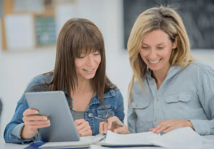 Two women smile while studying together, possibly taking general education courses online. One holds a tablet, the other a pen.