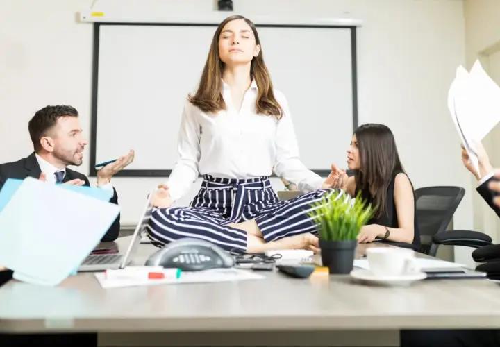 A woman meditates in lotus pose at a conference table while colleagues argue heatedly around her, creating a stark contrast between chaos and calm conversation.
