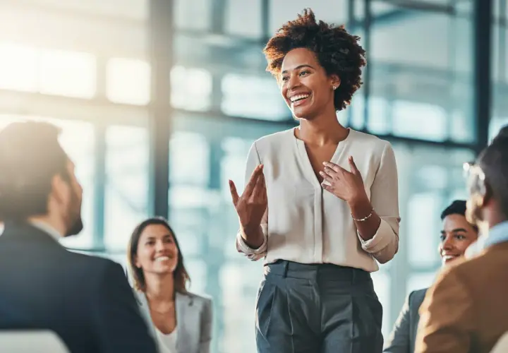 A woman leads a manager training session, engaging her audience with expressive gestures and a bright smile.