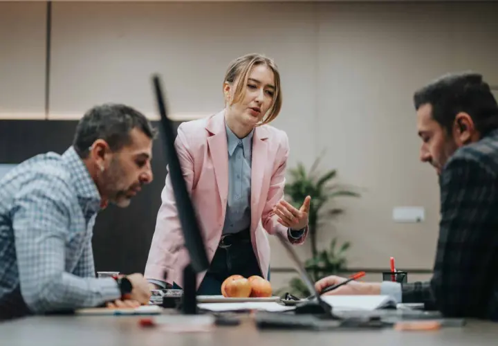 A woman in a pink blazer leads a discussion, demonstrating effective leadership and mentoring to her colleagues at a conference table.
