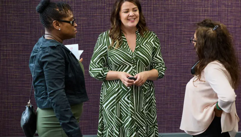 Three women in conversation, one in a green patterned dress, discussing strategies for driving performance.