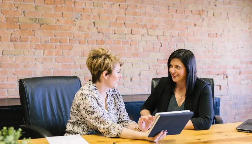 Two women in a meeting, one holding a tablet, with a brick wall background.