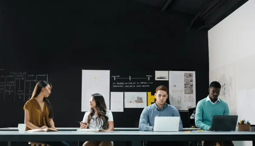 Four diverse individuals engage in a front line manager training session, collaborating around a table with laptops and notebooks.