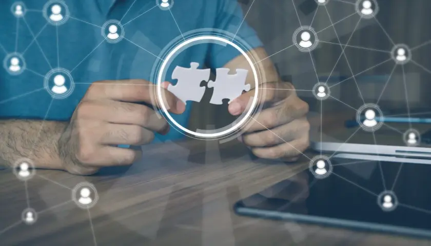 Hands connect puzzle pieces, symbolizing collaboration and leading a remote team through a network of connected people icons.