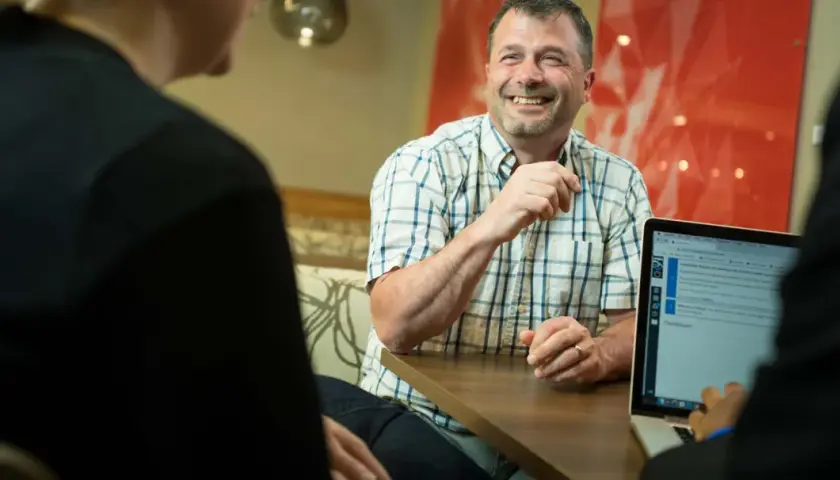 A man smiles while discussing his **associate degree in business online** with colleagues, looking at a laptop screen.