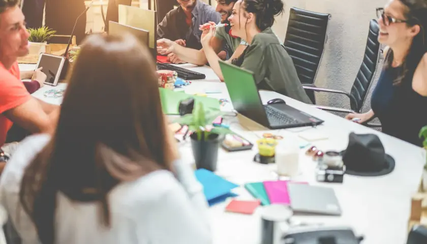 Millennials in the workplace collaborate around a table, sharing ideas and laughter. Laptops, tablets, and colorful notebooks suggest a dynamic, modern office environment.