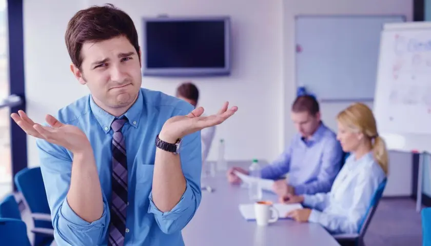 Confused man shrugs, wondering how to run meetings effectively while colleagues discuss in the background.