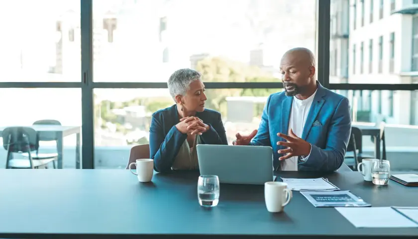 Two professionals discuss ideas for an internal communications podcast, leaning over a laptop at a conference table.