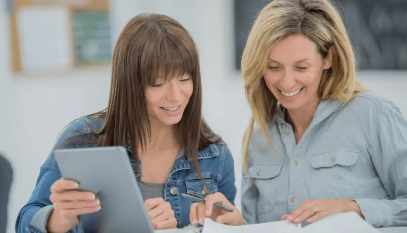 Two women smile while studying together, possibly taking general education courses online. One holds a tablet, the other a pen.
