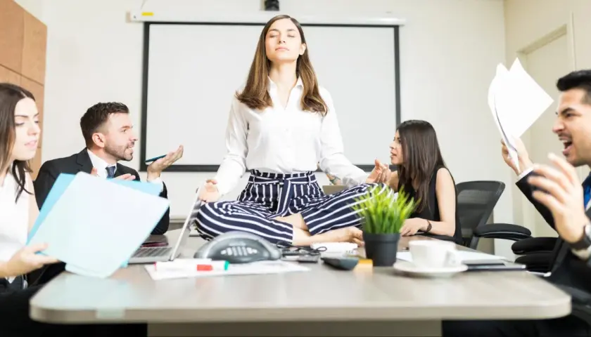 A woman meditates in lotus pose at a conference table while colleagues argue heatedly around her, creating a stark contrast between chaos and calm conversation.