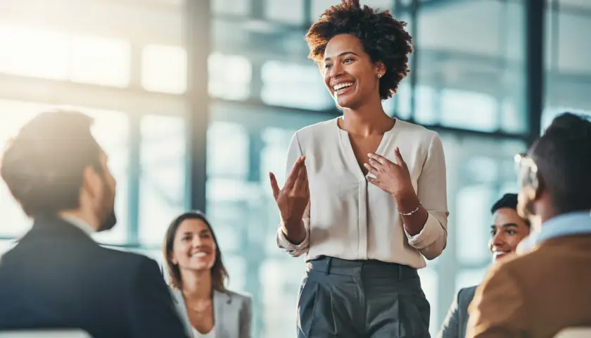 A woman leads a manager training session, engaging her audience with expressive gestures and a bright smile.