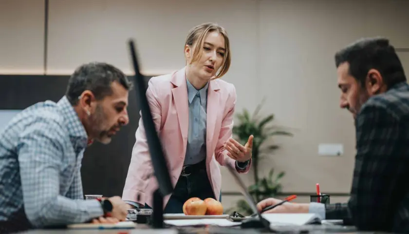 A woman in a pink blazer leads a discussion, demonstrating effective leadership and mentoring to her colleagues at a conference table.