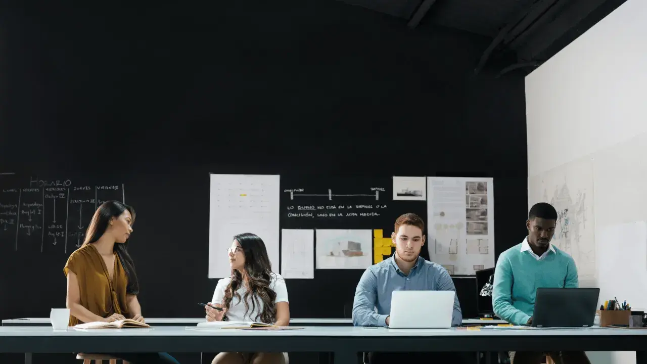 Four diverse individuals engage in a front line manager training session, collaborating around a table with laptops and notebooks.
