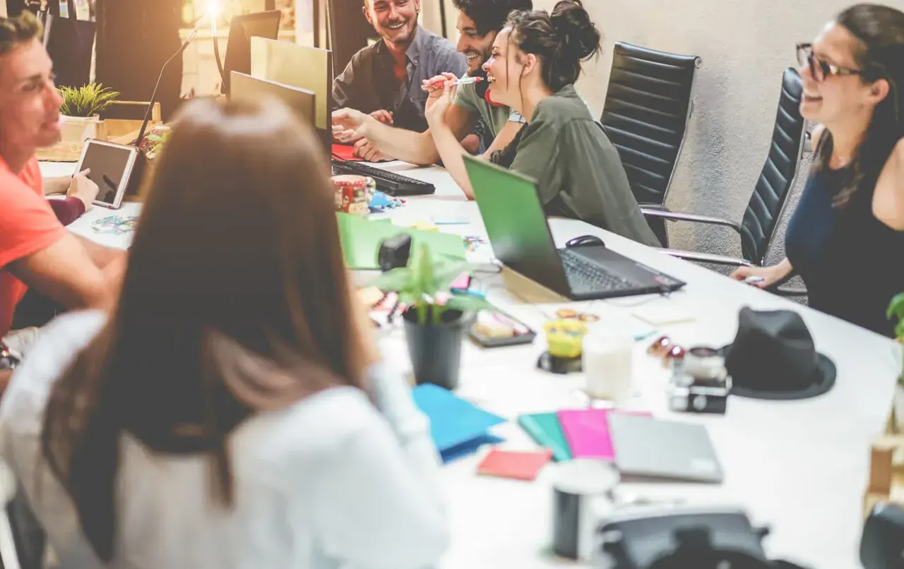 Millennials in the workplace collaborate around a table, sharing ideas and laughter. Laptops, tablets, and colorful notebooks suggest a dynamic, modern office environment.