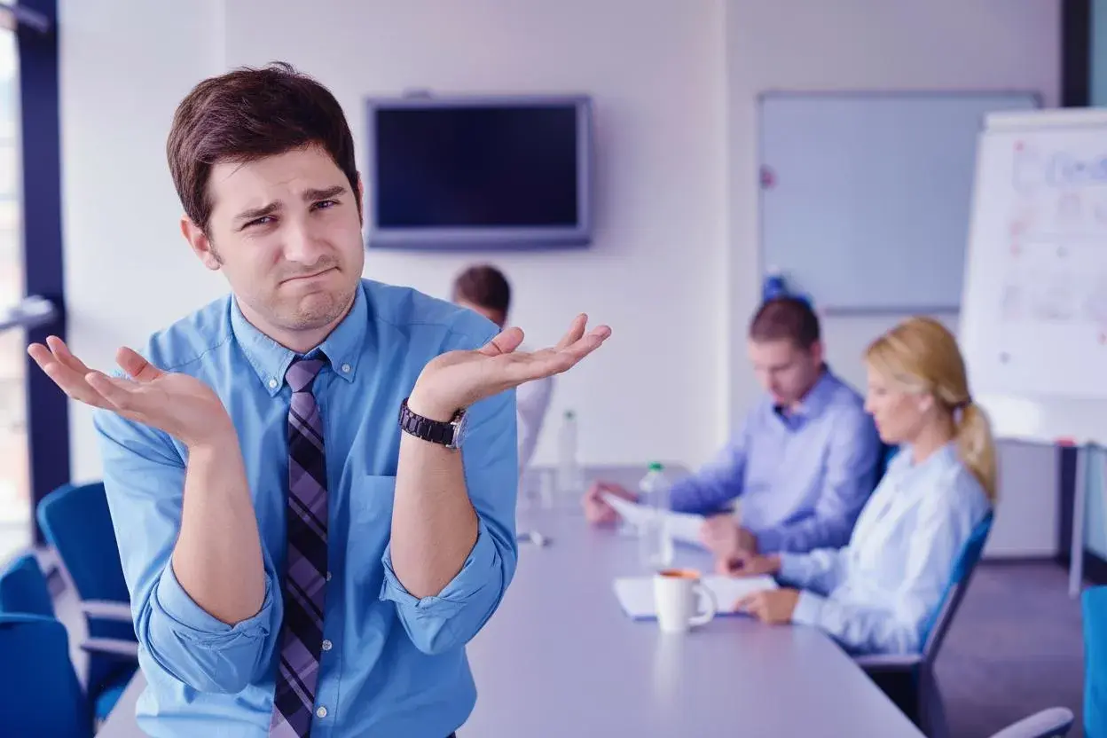 Confused man shrugs, wondering how to run meetings effectively while colleagues discuss in the background.