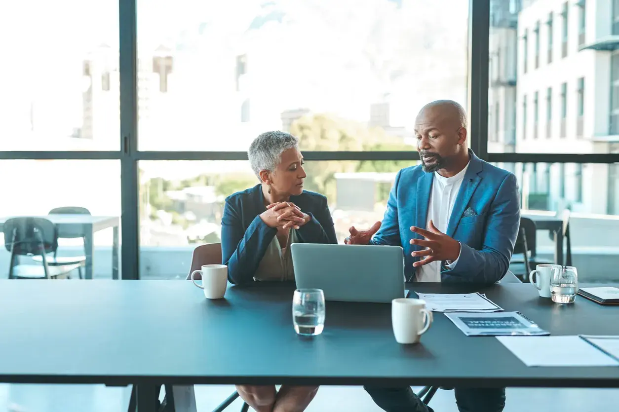 Two professionals discuss ideas for an internal communications podcast, leaning over a laptop at a conference table.