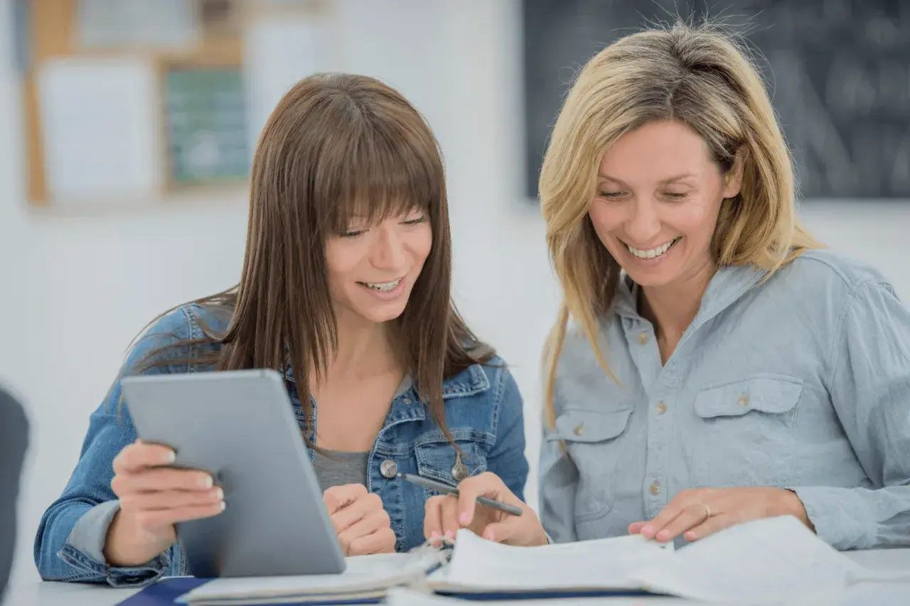Two women smile while studying together, possibly taking general education courses online. One holds a tablet, the other a pen.