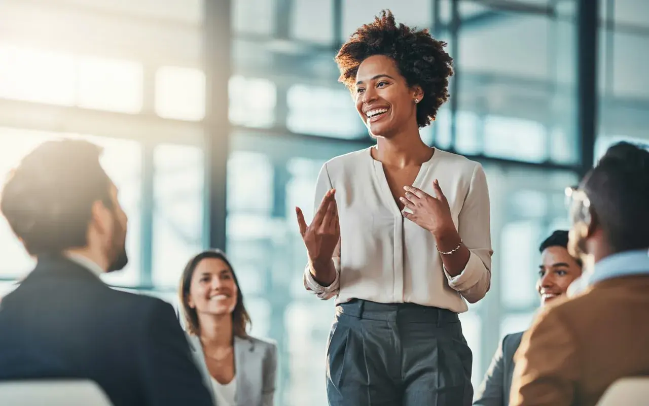 A woman leads a manager training session, engaging her audience with expressive gestures and a bright smile.