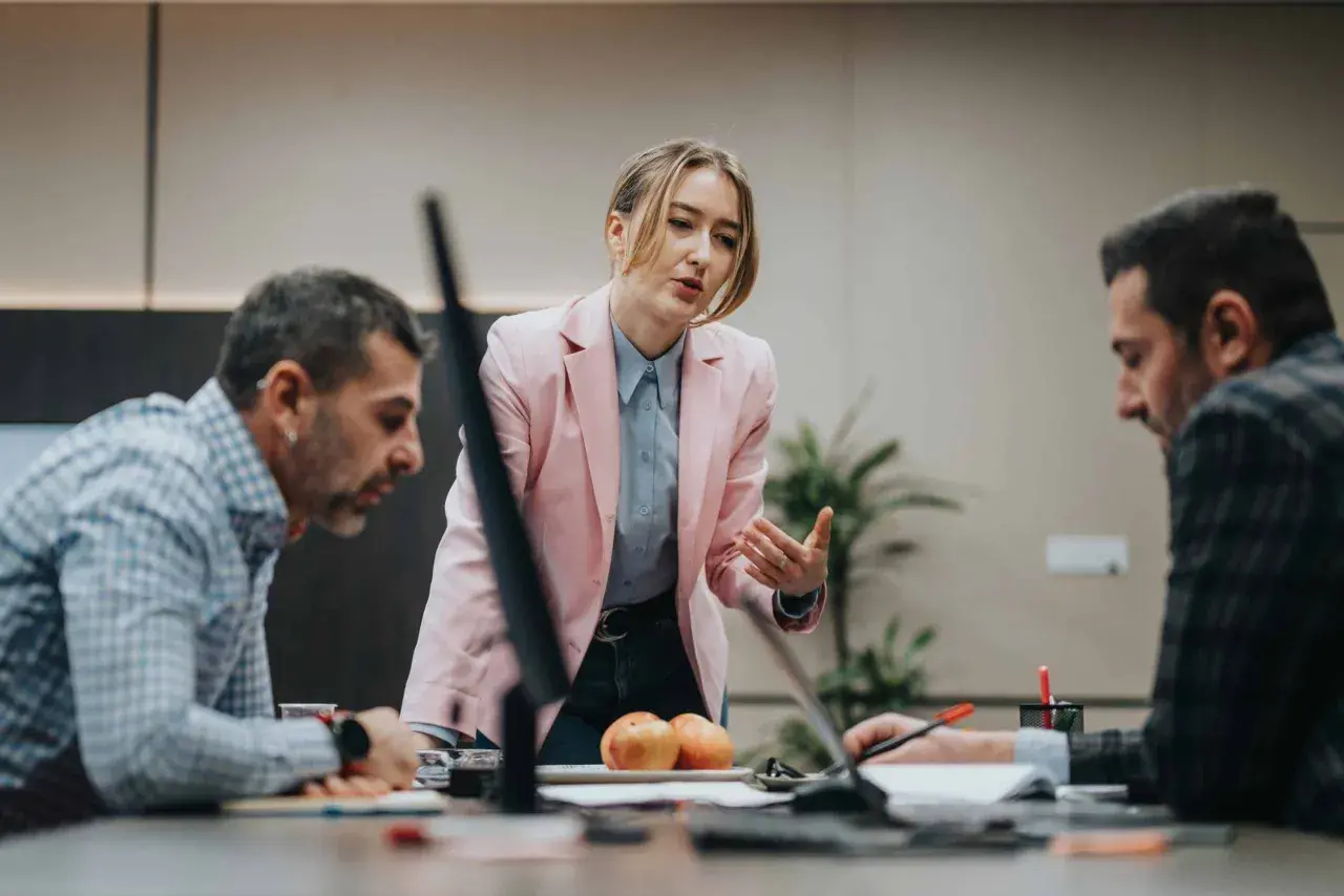 A woman in a pink blazer leads a discussion, demonstrating effective leadership and mentoring to her colleagues at a conference table.