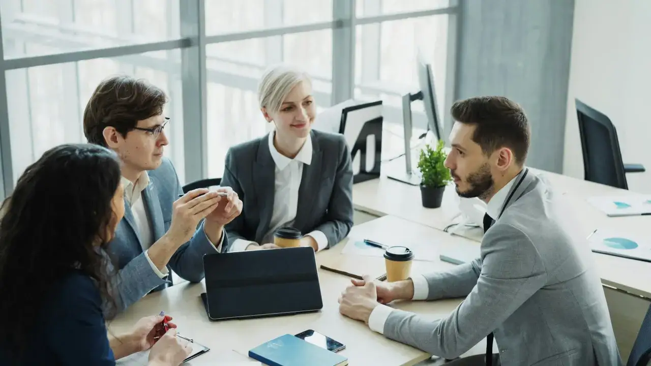 A collaborative team of four professionals in a modern office, discussing ideas around a table with a tablet and coffee cups.