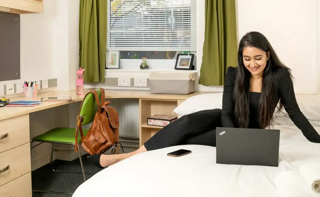 A young woman smiles while working on her laptop, likely pursuing an online associate degree program from her dorm room.