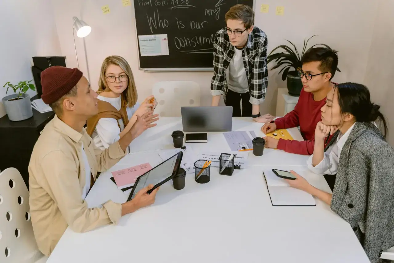 A diverse team collaborates around a table, discussing strategy. The whiteboard reads 