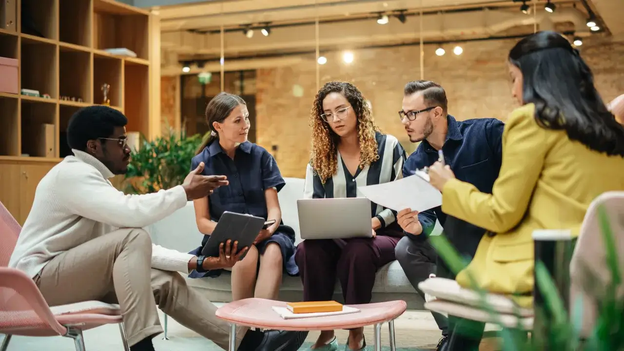 A diverse team collaborates, showcasing influential leadership through active discussion and shared focus on a laptop and documents.