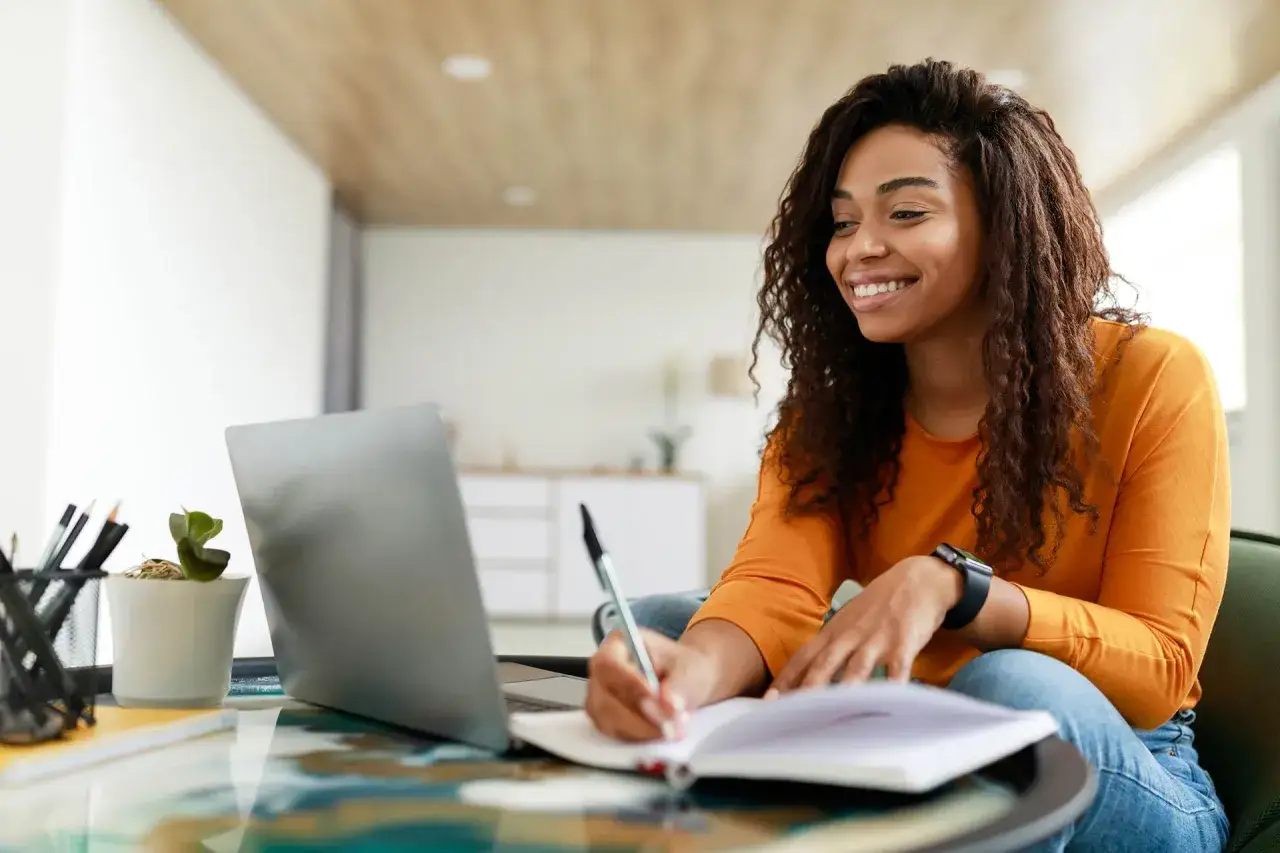 A smiling woman studies computer information systems online, taking notes on her laptop and in a notebook.