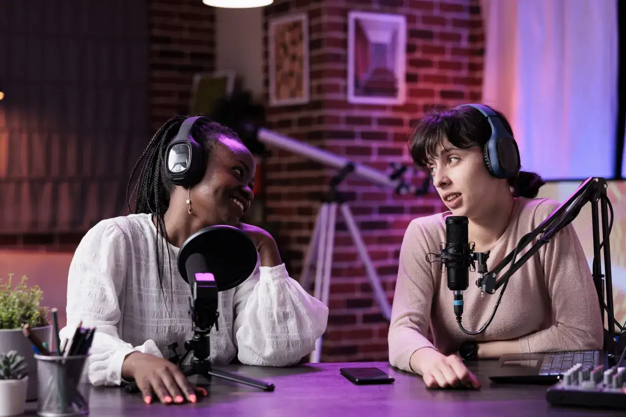 Two women with headphones record internal company podcasts, smiling and engaging with microphones in a studio setting.
