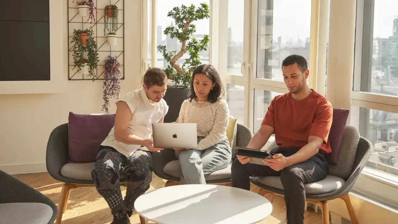 Three students collaborate on a laptop, perhaps studying for an associate business degree online, in a bright room with city views.