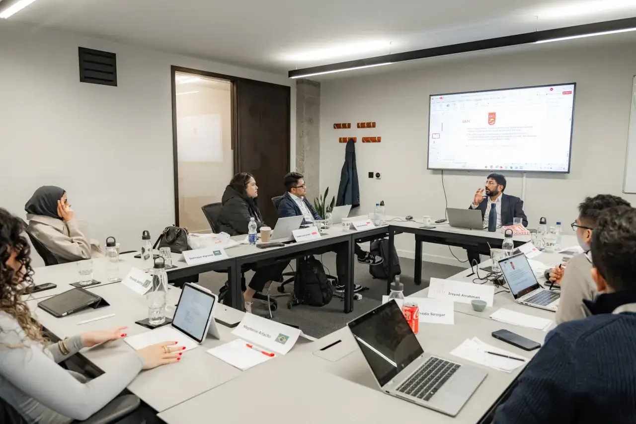 A diverse group engages in a leadership coaching session around a conference table, with laptops and notes suggesting active learning.
