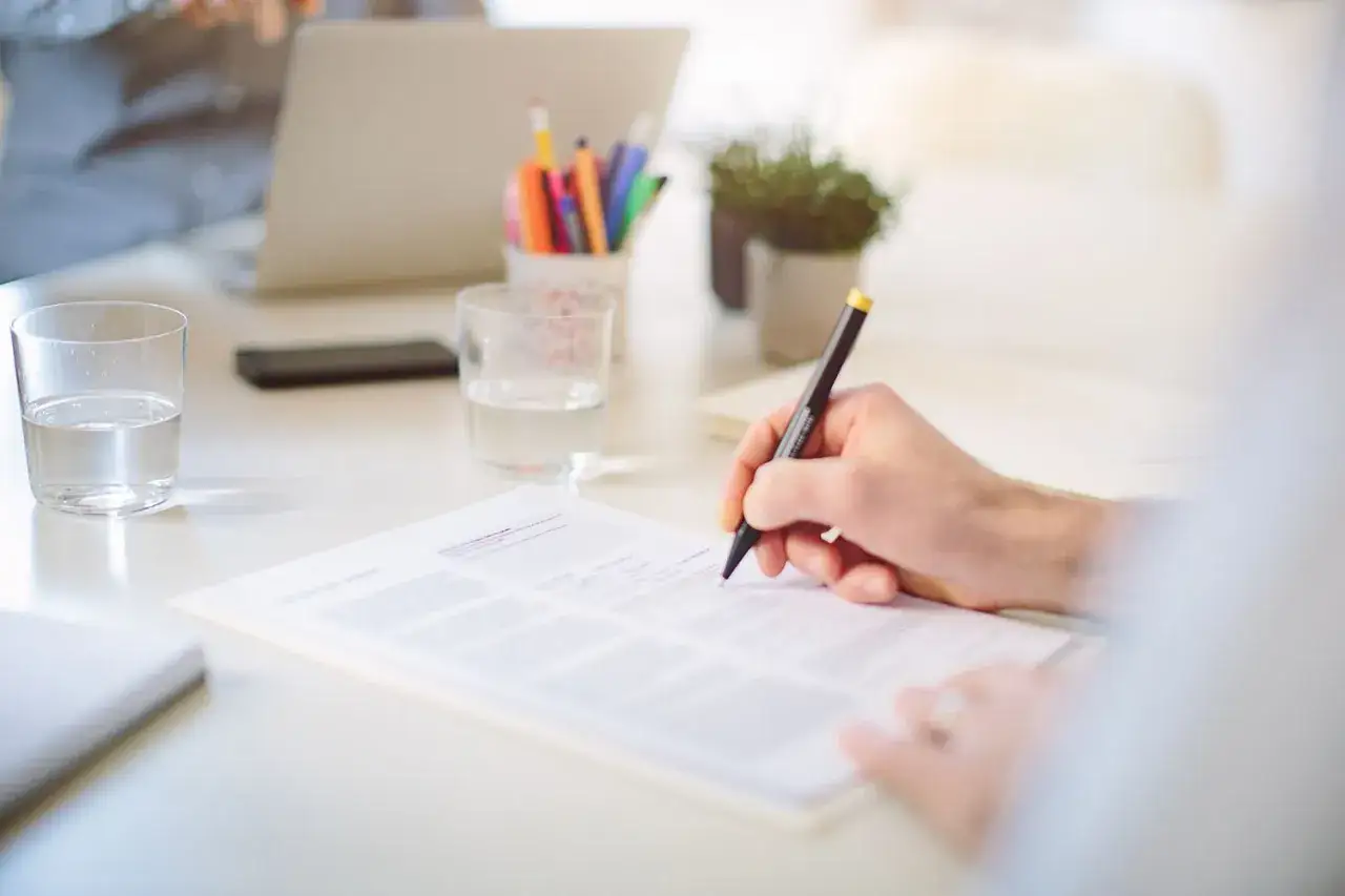 A hand writes on a document, likely a cover letter, at a desk with glasses of water, pens, and a laptop.