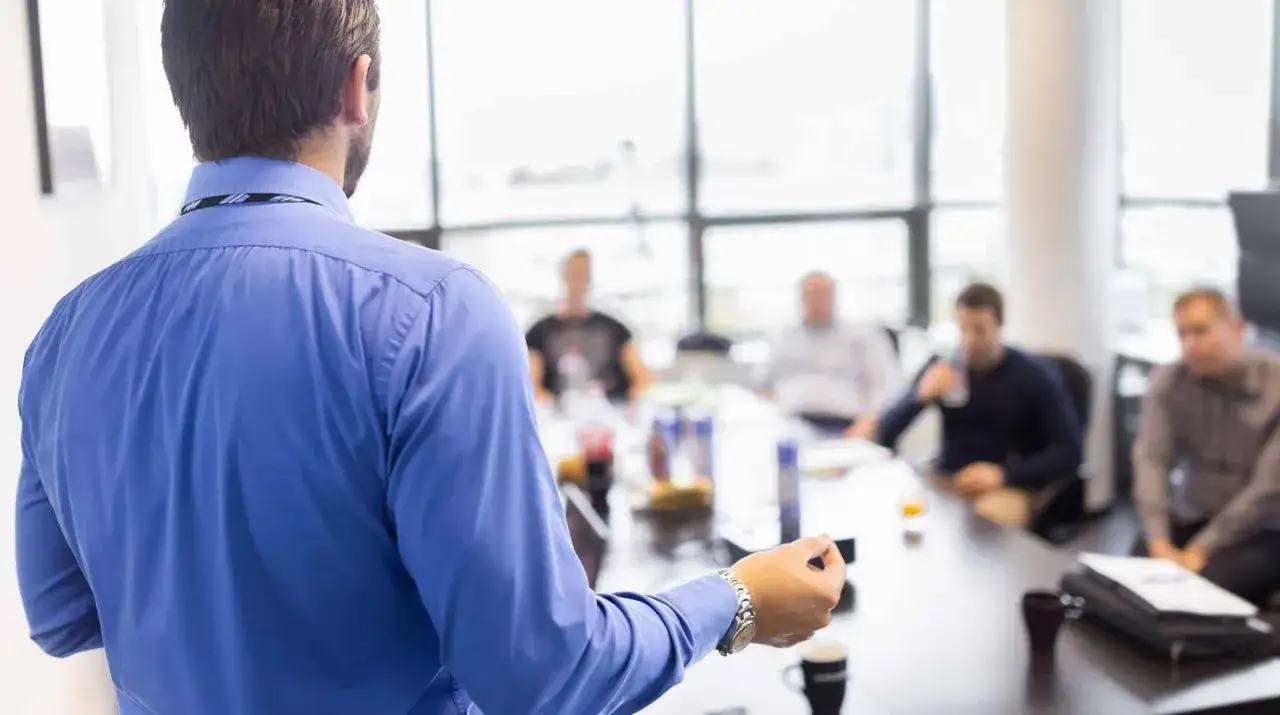 A man in a blue shirt leads a delegation training session, gesturing to a group seated around a conference table.